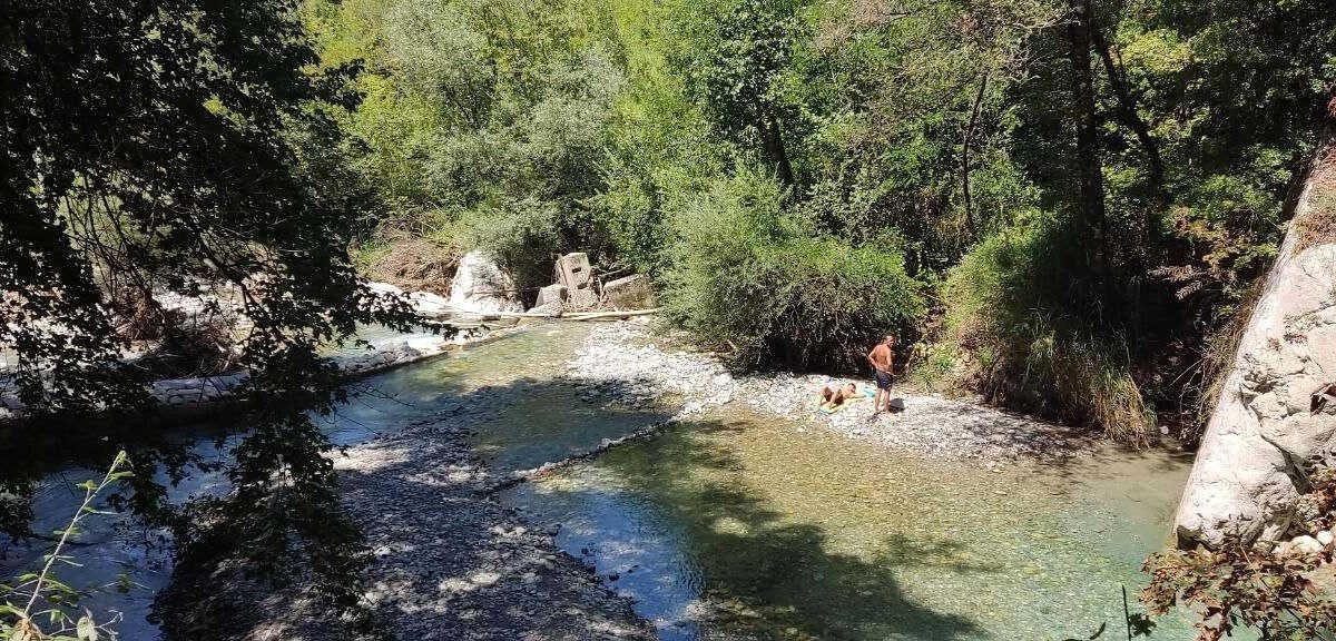 Riva di un fiume immersa in natura, con persone che si rilassano e si godono l'acqua cristallina in un ambiente verde e suggestivo.