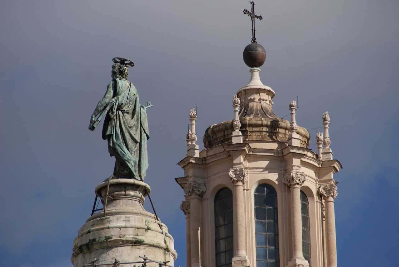Colonna Traiana un film di pietra in 155 scene 3 Santuario sul tetto di un edificio storico con una statua di un santo e una coppa decorativa con croce, simbolo religioso in stile barocco, cielo nuvoloso sullo sfondo, architettura religiosa.
