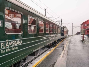 Treno storico sulla Ferrovia Flåm in Norvegia con paesaggi montani innevati, situazione di sosta alla stazione di Flåm, atmosfere autunnali e viaggi panoramici in Norvegia.