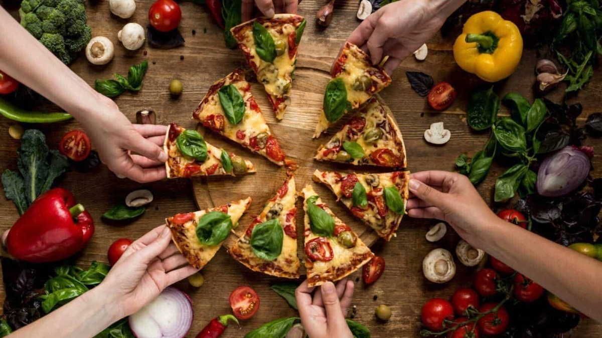 Fantasiosa pizza margherita con basilico, pomodori e formaggio, afferrata da diverse mani, su tavolo di legno con verdure fresche.