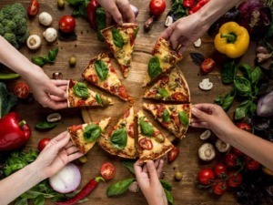 Fantasiosa pizza margherita con basilico, pomodori e formaggio, afferrata da diverse mani, su tavolo di legno con verdure fresche.