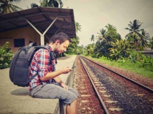 Viaggiatore giovane con zaino che controlla il telefono alla stazione ferroviaria in una zona tropicale, ambientato in un contesto di avventura e turismo sostenibile.