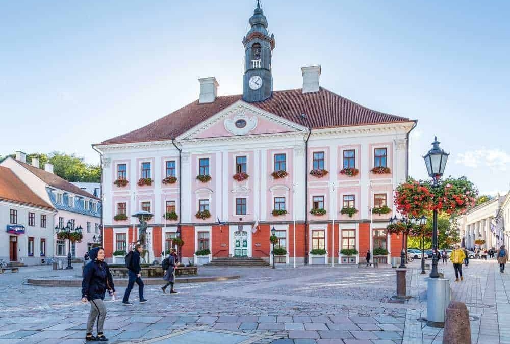 Villa storica nel centro storico di una città europea, edificio con facciata rosa e dettagli decorativi in bianco, torre con orologio, piazza vivace con persone e lampioni storici.
