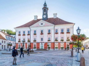 Villa storica nel centro storico di una città europea, edificio con facciata rosa e dettagli decorativi in bianco, torre con orologio, piazza vivace con persone e lampioni storici.