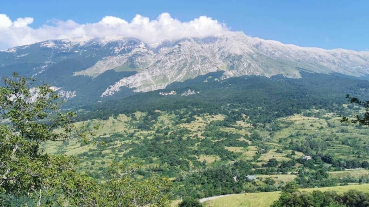 Montagna imponente con cime rocciose e vegliata da nuvole, circondata da verdi colline e foreste rigogliose, rappresentando un paesaggio naturale mozzafiato in Italia.