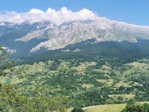 Montagna imponente con cime rocciose e vegliata da nuvole, circondata da verdi colline e foreste rigogliose, rappresentando un paesaggio naturale mozzafiato in Italia.