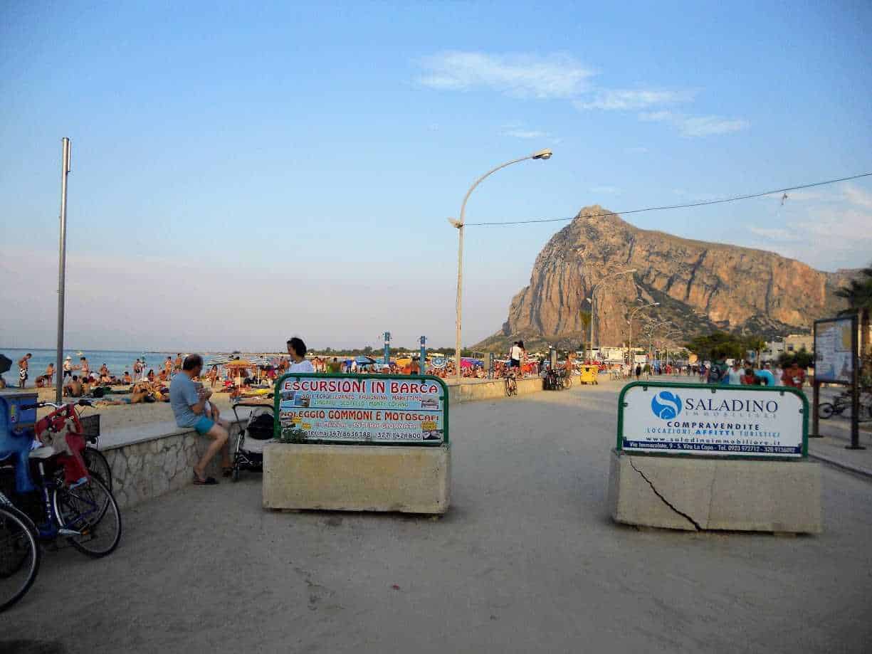 Spiaggia di San Vito Lo Capo, con sabbia dorata e mare cristallino, bagnanti e montagne sullo sfondo, in una giornata estiva soleggiata.