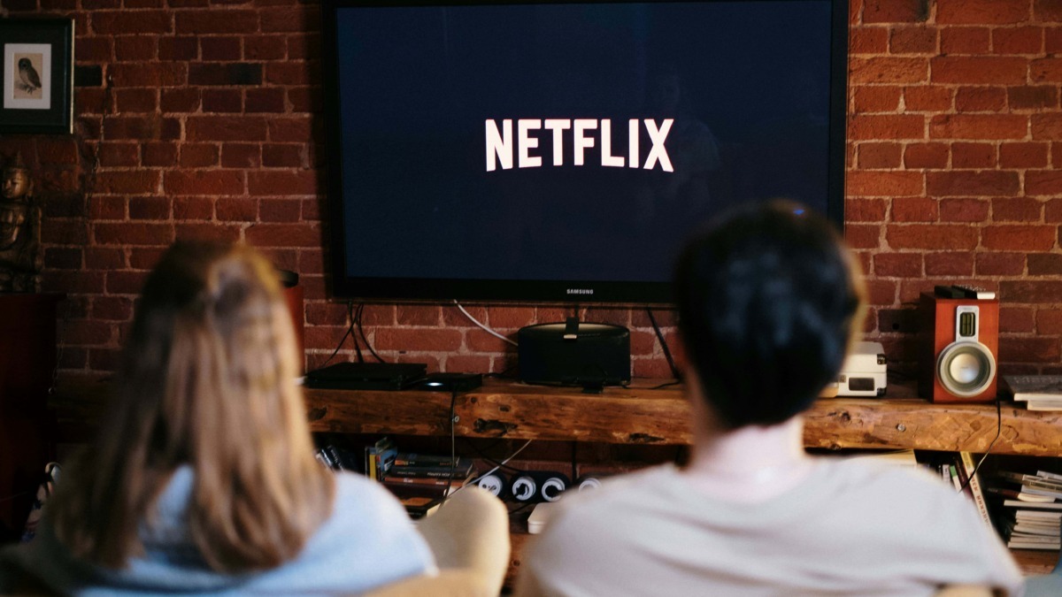 Schermo TV con logo Netflix che mostra una scena di visione di due persone sedute su un divano, ambiente domestico con pareti in mattoni rossi e arredamento vintage.