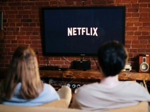 Schermo TV con logo Netflix che mostra una scena di visione di due persone sedute su un divano, ambiente domestico con pareti in mattoni rossi e arredamento vintage.