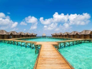 Overwater bungalows in a tropical resort with clear turquoise water and a wooden pier under a blue sky with fluffy clouds.