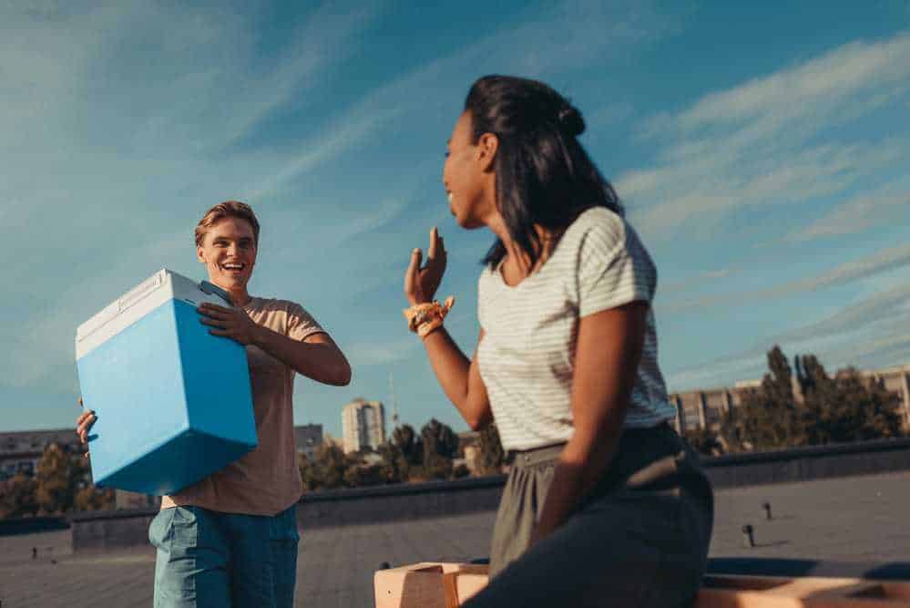 Titolare di blog, ragazza felice dai capelli scuri conversa con giovane che consegna un pacco azzurro, ambiente urbano al tramonto, atmosfera di amicizia e networking, immagini di consegna pacchi e social media.