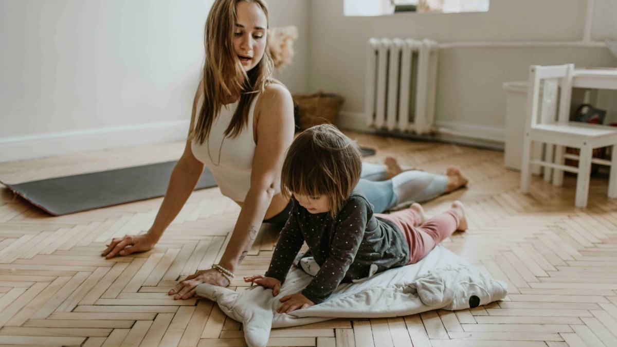 Altezza di donna e bambina che praticano stretching o yoga in casa, su un pavimento in parquet, con luce naturale, ambiente domestico rilassato e accogliente.
