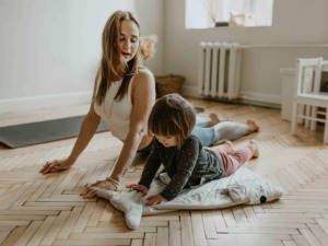 Altezza di donna e bambina che praticano stretching o yoga in casa, su un pavimento in parquet, con luce naturale, ambiente domestico rilassato e accogliente.