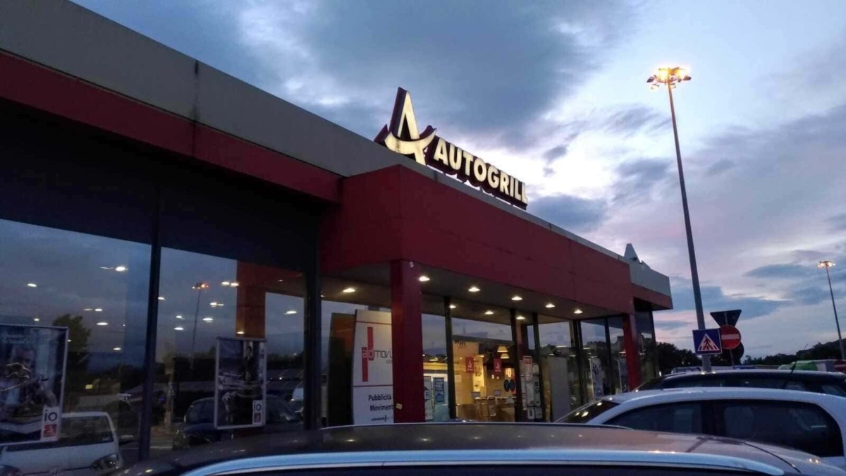 Autogrill, ristorante e area di servizio presso autostrada, edificio moderno con grandi vetrate, parcheggio e insegne luminose, atmosfera serale con cielo nuvoloso, illuminazione esterna.