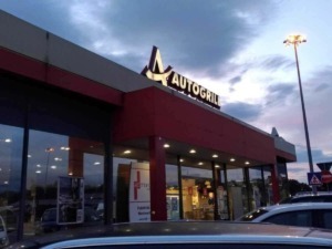 Autogrill, ristorante e area di servizio presso autostrada, edificio moderno con grandi vetrate, parcheggio e insegne luminose, atmosfera serale con cielo nuvoloso, illuminazione esterna.