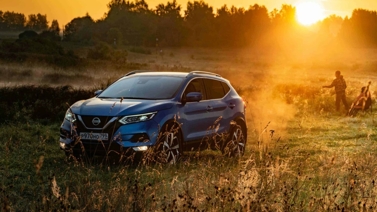 Nissan Qashqai blu accanto a un campo al tramonto con persone che giocano, ambientazione naturale, luce calda e atmosfera serena.