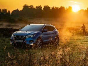 Nissan Qashqai blu accanto a un campo al tramonto con persone che giocano, ambientazione naturale, luce calda e atmosfera serena.