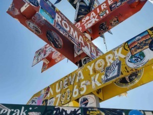 Cartelli stradali ricoperti di adesivi e graffiti, segnavia di direzioni e mappe, simboli e testo, presso una località turistica o di interesse, con cielo blu sullo sfondo.