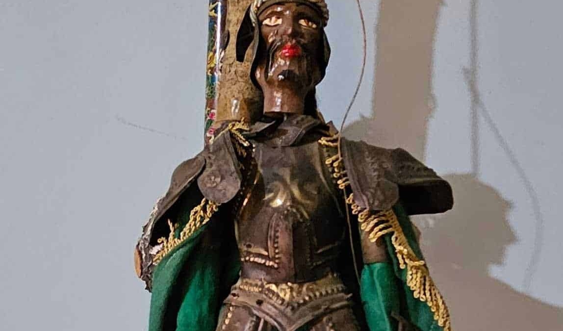 Antico statua in legno di una figura africana tradizionale vestita con abiti decorativi, posta contro un muro in un ambiente interno, rappresentando l'arte e la cultura africana.