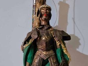 Antico statua in legno di una figura africana tradizionale vestita con abiti decorativi, posta contro un muro in un ambiente interno, rappresentando l'arte e la cultura africana.