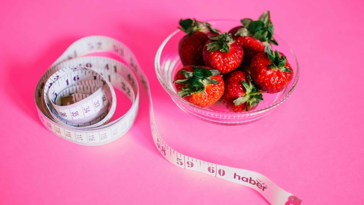 Fette di plastica e ciotola di fragole fresche con nastro da misurazione su sfondo rosa.