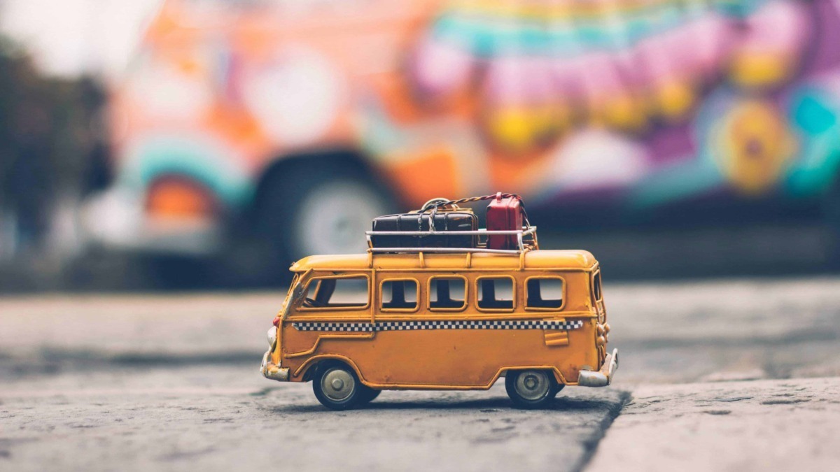 Mini camper van giocattolo arancione con valigie sul tetto, su strada di pietra, con macchina colorata sfocata sullo sfondo, stile rustico e vintage.
