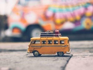 Mini camper van giocattolo arancione con valigie sul tetto, su strada di pietra, con macchina colorata sfocata sullo sfondo, stile rustico e vintage.