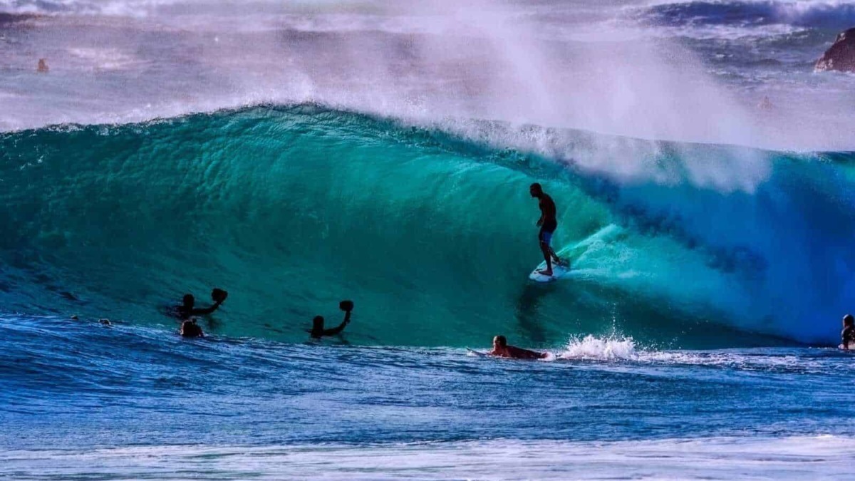 Surfista che cavalca un'onda gigante in mare, con altri surfisti che lo supportano, acque cristalline e onde maestose, scena di surf e sport acquatici, atmosfera estiva e avventurosa.