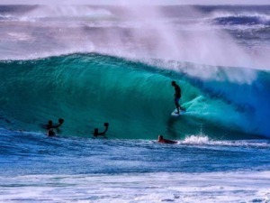 Surfista che cavalca un'onda gigante in mare, con altri surfisti che lo supportano, acque cristalline e onde maestose, scena di surf e sport acquatici, atmosfera estiva e avventurosa.