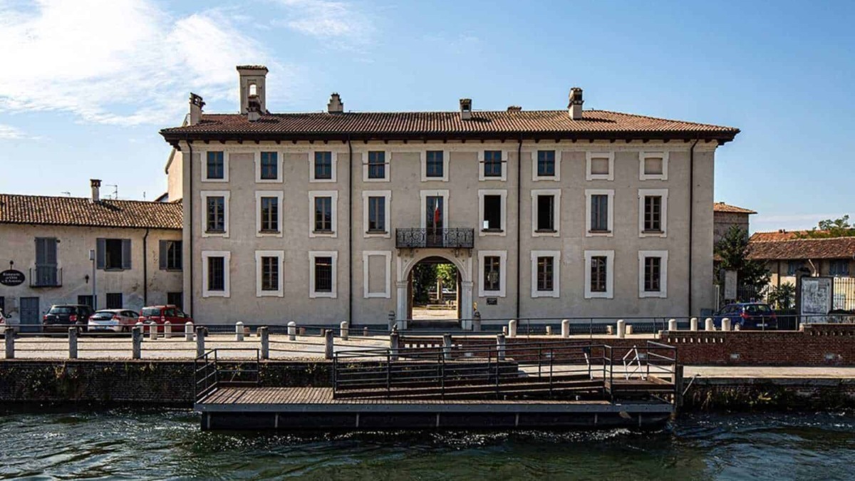 Palazzo storico sulla riva del fiume con ingresso ad arco, situato in un villaggio pittoresco, con auto parcheggiate e un paesaggio urbano suggestivo, ideale per fotografie di architettura e turismo.