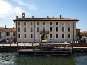 Palazzo storico sulla riva del fiume con ingresso ad arco, situato in un villaggio pittoresco, con auto parcheggiate e un paesaggio urbano suggestivo, ideale per fotografie di architettura e turismo.