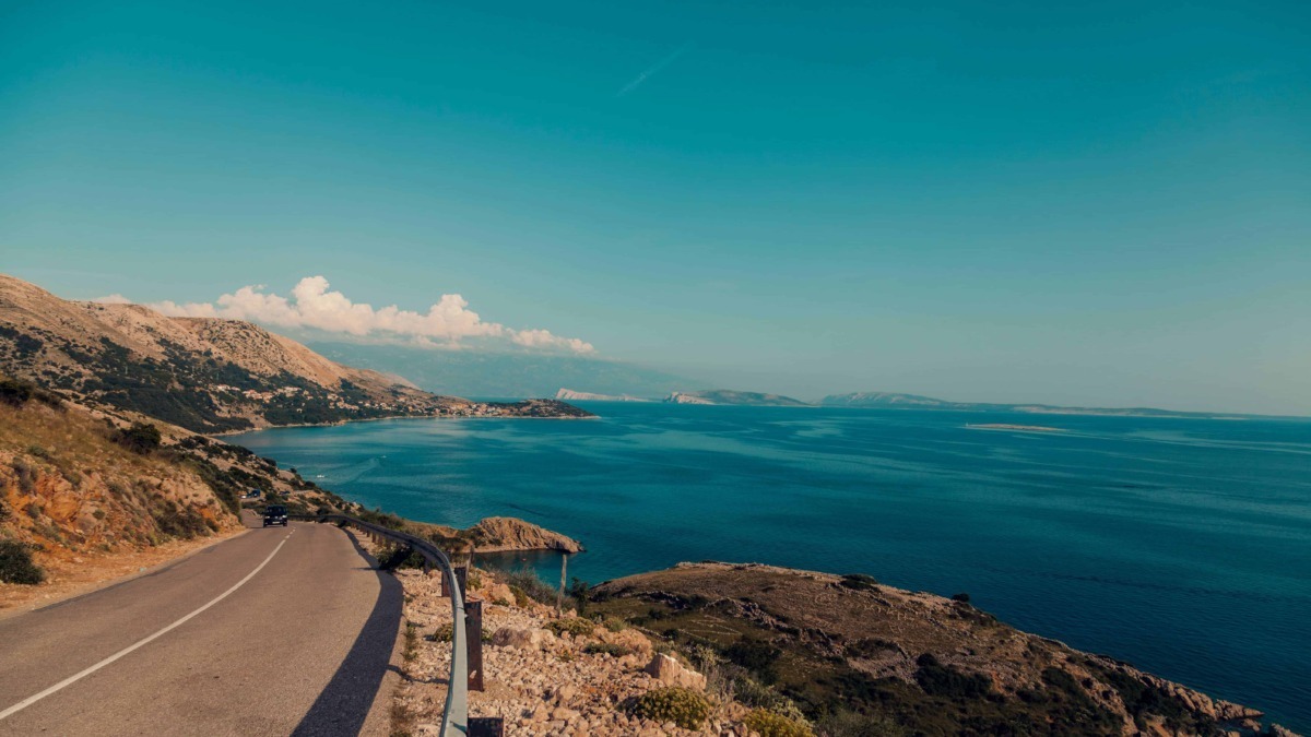 Tragitto costiero panoramico con vista mare e colline, strada asfaltata con guardrail, paesaggio mediterraneo, viaggio in auto lungo la costa, bellezza naturale, scenari di mare e montagna in Italia.