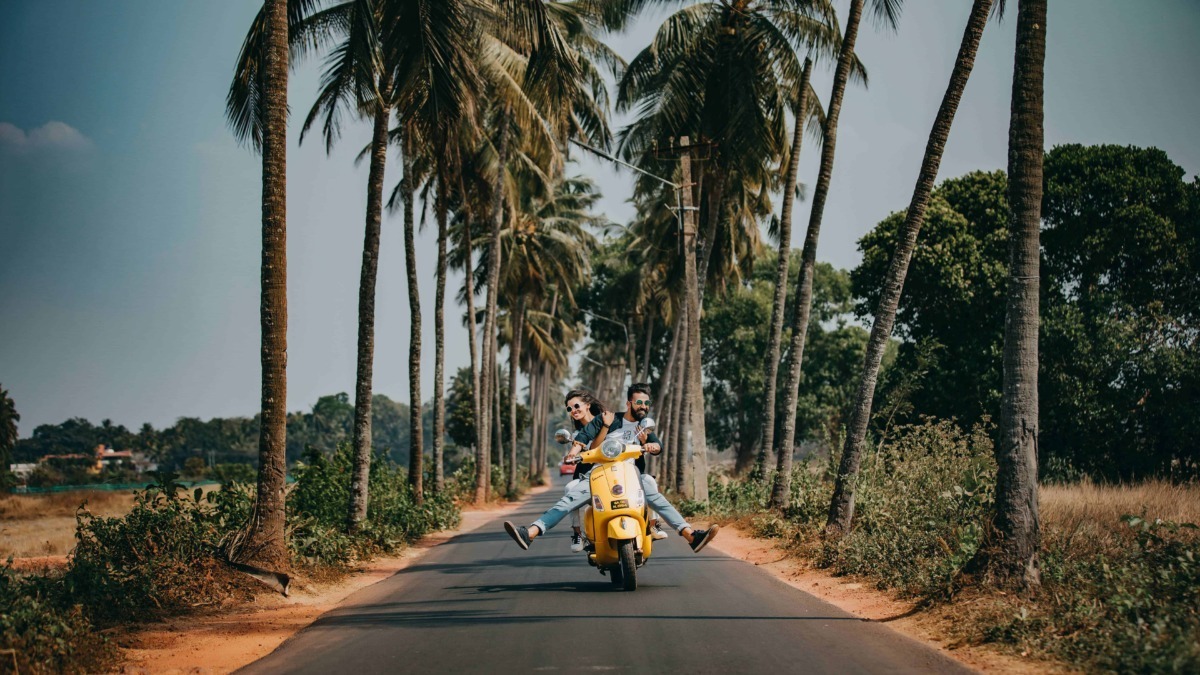 Motociclisti che ridono e fanno acrobazie su una Vespa gialla in una strada panoramica tra palme e natura tropicale in una località di vacanza.