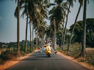 Motociclisti che ridono e fanno acrobazie su una Vespa gialla in una strada panoramica tra palme e natura tropicale in una località di vacanza.