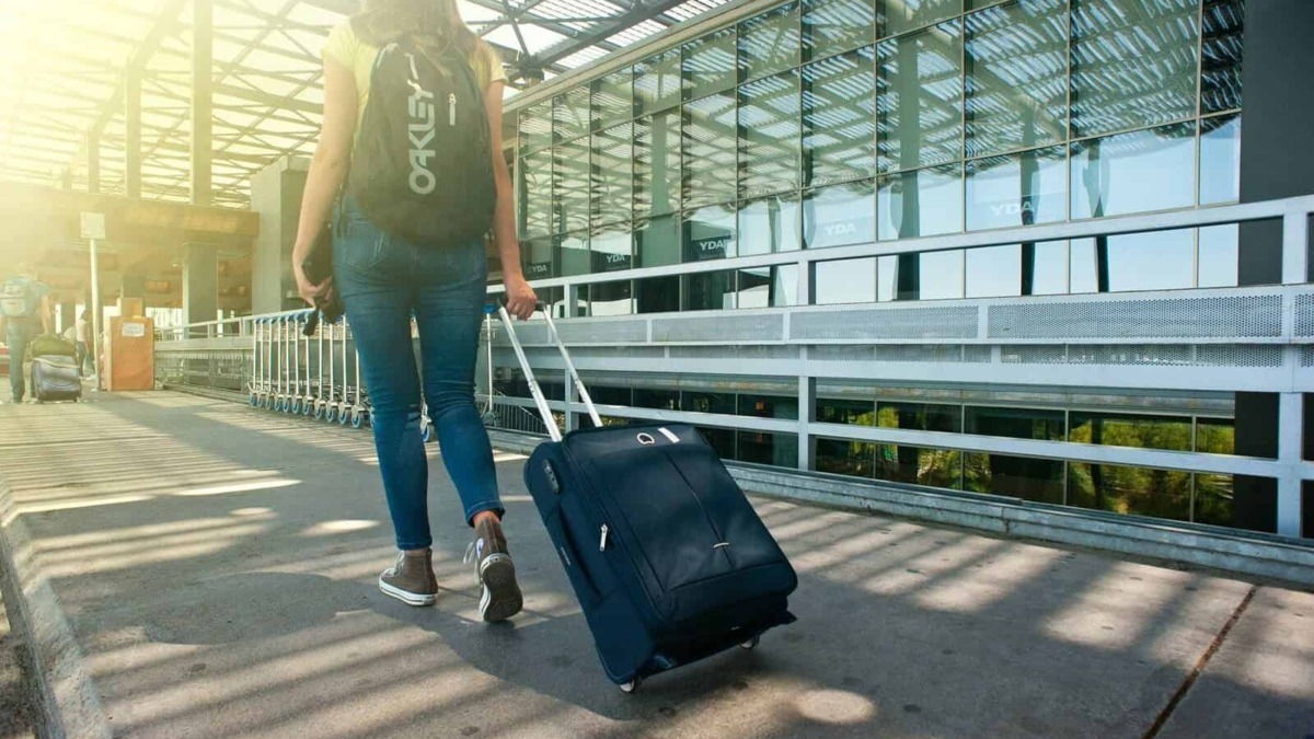 Bagaglio a mano con rotelle all'aeroporto, giovane donna con zaino in spalla e trolley in mano, viaggio e turismo, moderna struttura aeroportuale in background.