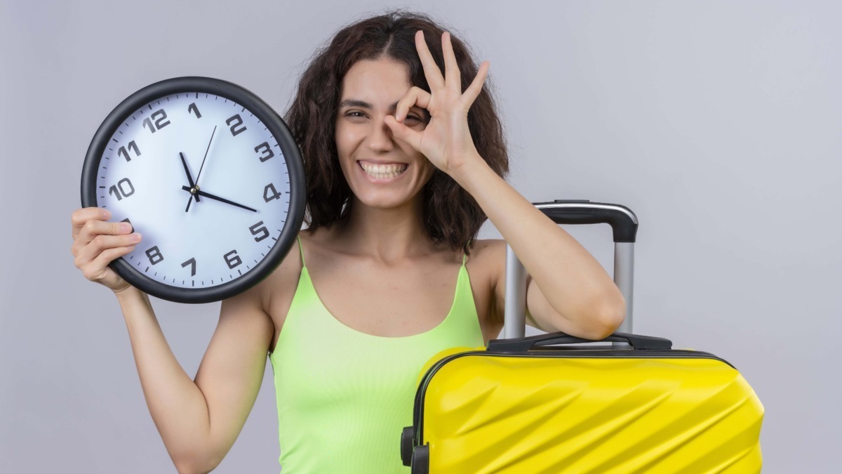 Orologio grande con lancette che segnano le 12:13 in mano a una donna sorridente con bagaglio giallo e gesto occhi a forma di occhio.
