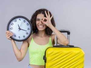 Orologio grande con lancette che segnano le 12:13 in mano a una donna sorridente con bagaglio giallo e gesto occhi a forma di occhio.