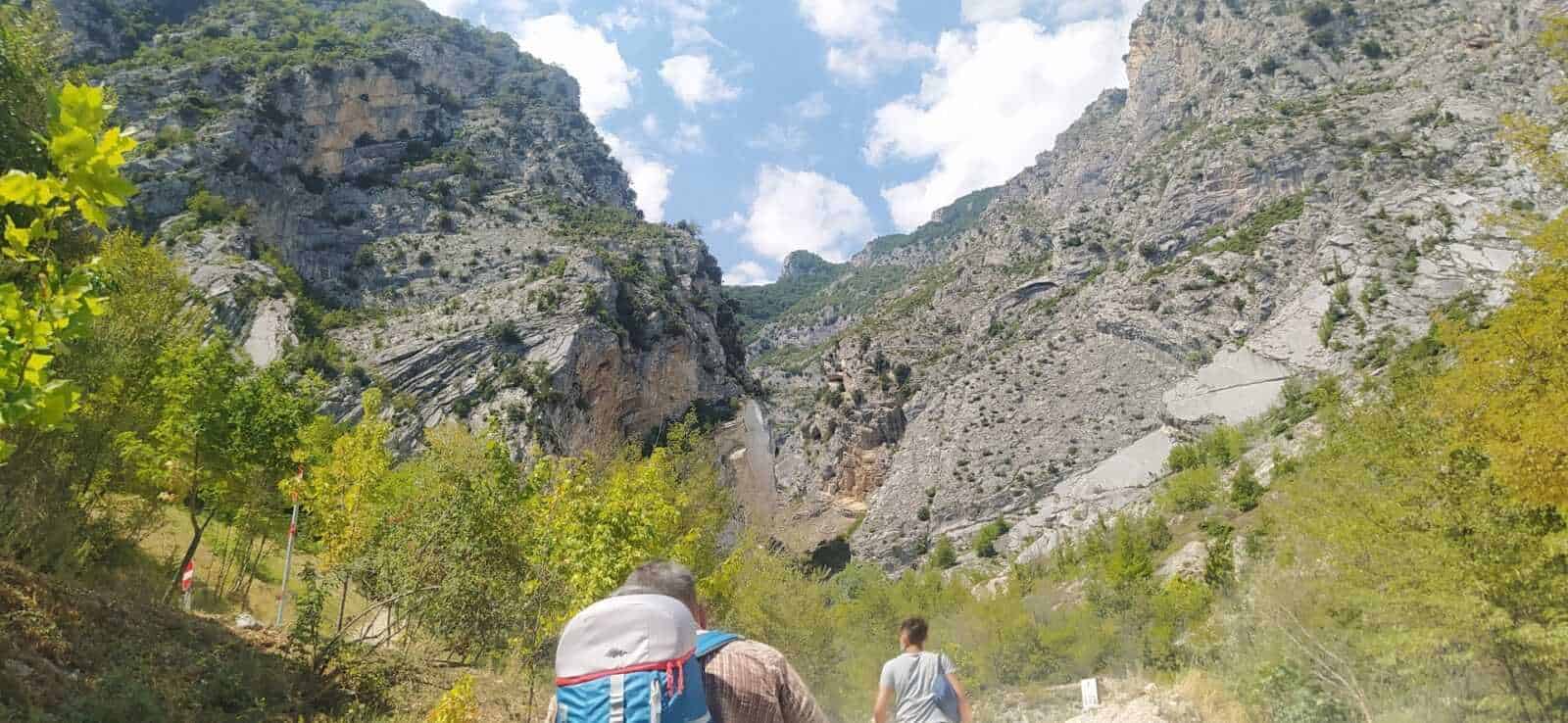 Escursionisti in montagna con zaini che attraversano una valle alpina circondata da scoscese pareti rocciose e vegetazione verde, in un paesaggio naturale e suggestivo.