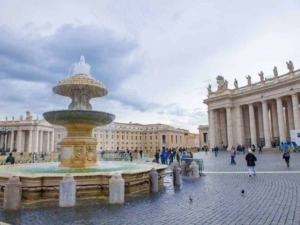 Fontana storica in piazza San Pietro a Roma, con colonne e statue, visitatori e cielo nuvoloso, simbolo di arte e cultura italiana, ideale per blog di viaggi e turismo.