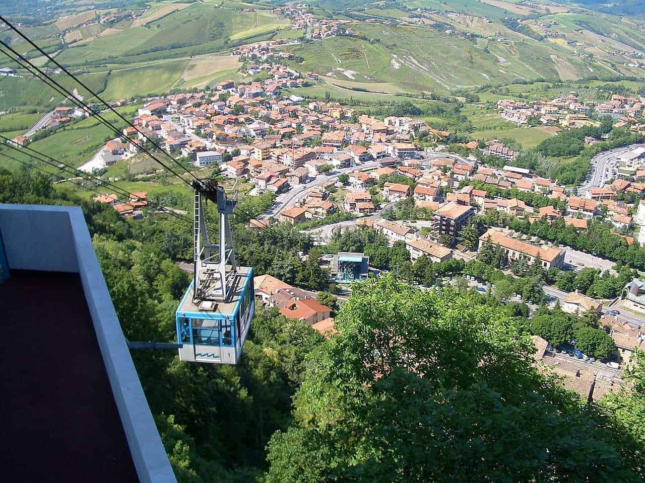 San Marino weekend estivo montagna 7 Vettura funicolare che si arrampica sulla collina con vista panoramica di un borgo italiano tra verdeggianti colline, case con tetti di terracotta e strade sinuose, immerso in una natura rigogliosa.