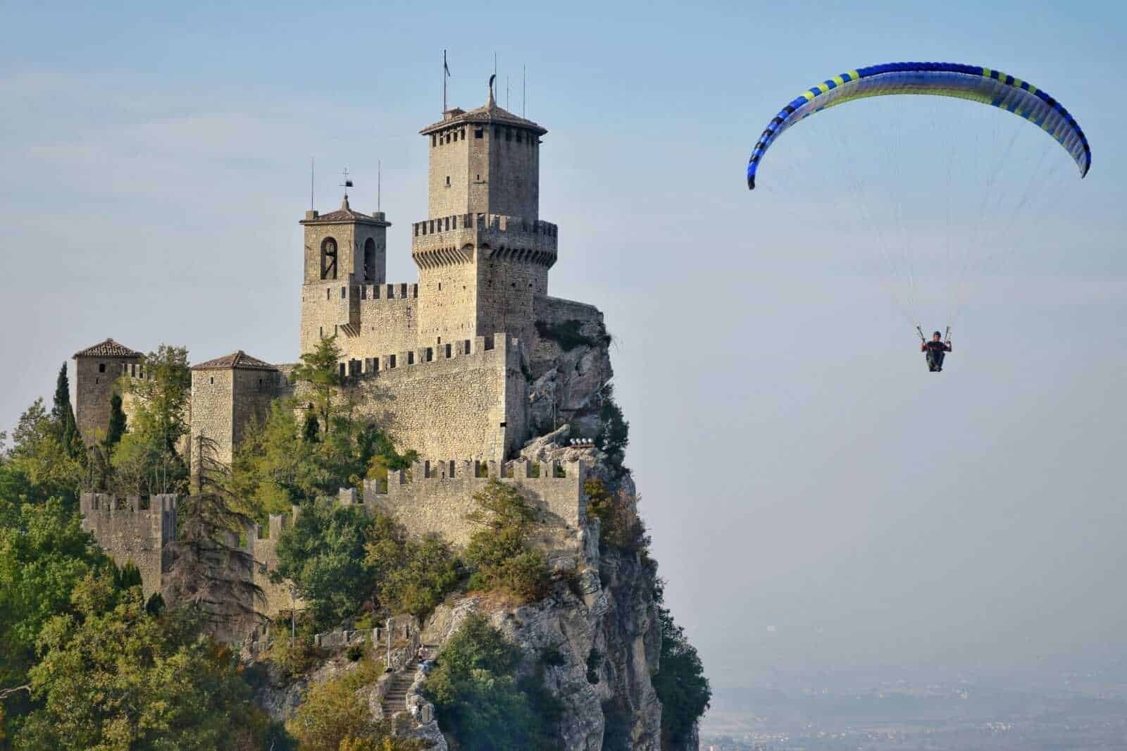 San Marino weekend estivo montagna 5 Castello sul vertiginoso scoglio con parachutista che atterra nei pressi, panoramica vista di Torre e mura medievali con paesaggio naturale e cielo sereno.