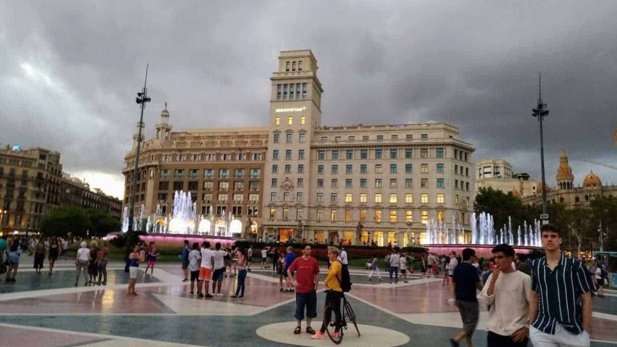 Fontana Plaça de Catalunya a Barcellona con persone e edificio storico in sfondo durante un tramonto nuvoloso, scena urbana vivace e attrazione turistica.