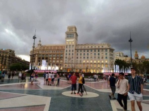Fontana Plaça de Catalunya a Barcellona con persone e edificio storico in sfondo durante un tramonto nuvoloso, scena urbana vivace e attrazione turistica.