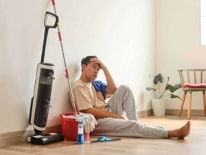 Spazzatura, aspirapolvere, e uomo stanco seduto sul pavimento in cucina, simbolo di affaticamento domestico e pulizie di casa, in un ambiente luminoso e minimalista.
