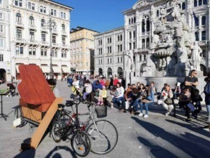 Sedie e persone che attendono in piazza con fontana storica, edifici antichi e cielo azzurro sullo sfondo, scena vivace di un centro urbano italiano.