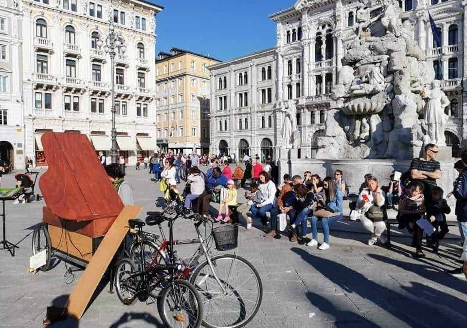 Sedie e persone che attendono in piazza con fontana storica, edifici antichi e cielo azzurro sullo sfondo, scena vivace di un centro urbano italiano.