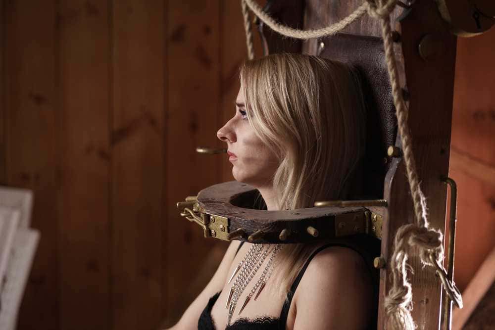 Collare di tortura medievale con donna seduta, immagine storica, ambiente in legno, versione moderna e originale, tema di torture e storia medievale, rappresentazione di tortura e sofferenza, contenuti storici e di interesse culturale, immagine di prova di resistenza e tormento.