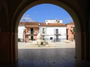 Villaggio di case colorate e piazza con fontana, vista attraverso un arco, in una tipica scena italiana.