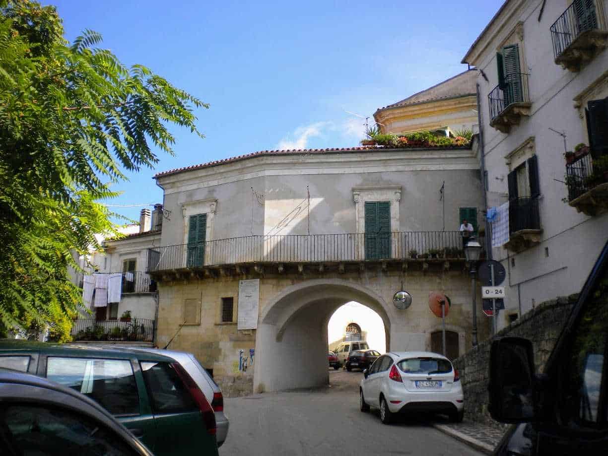 Architettura storica con arco di passaggio, edifici con balconi, auto parcheggiate, in un paesino italiano, cuore del centro storico, atmosfera mediterranea, strade pittoresche, quadro urbano autentico.