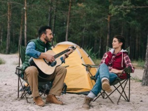 Un uomo e una donna seduti intorno a una tenda in natura, con l'uomo che suona la chitarra, ambiente boschivo e atmosfera rilassante, perfetto per campeggio e musica all'aperto.